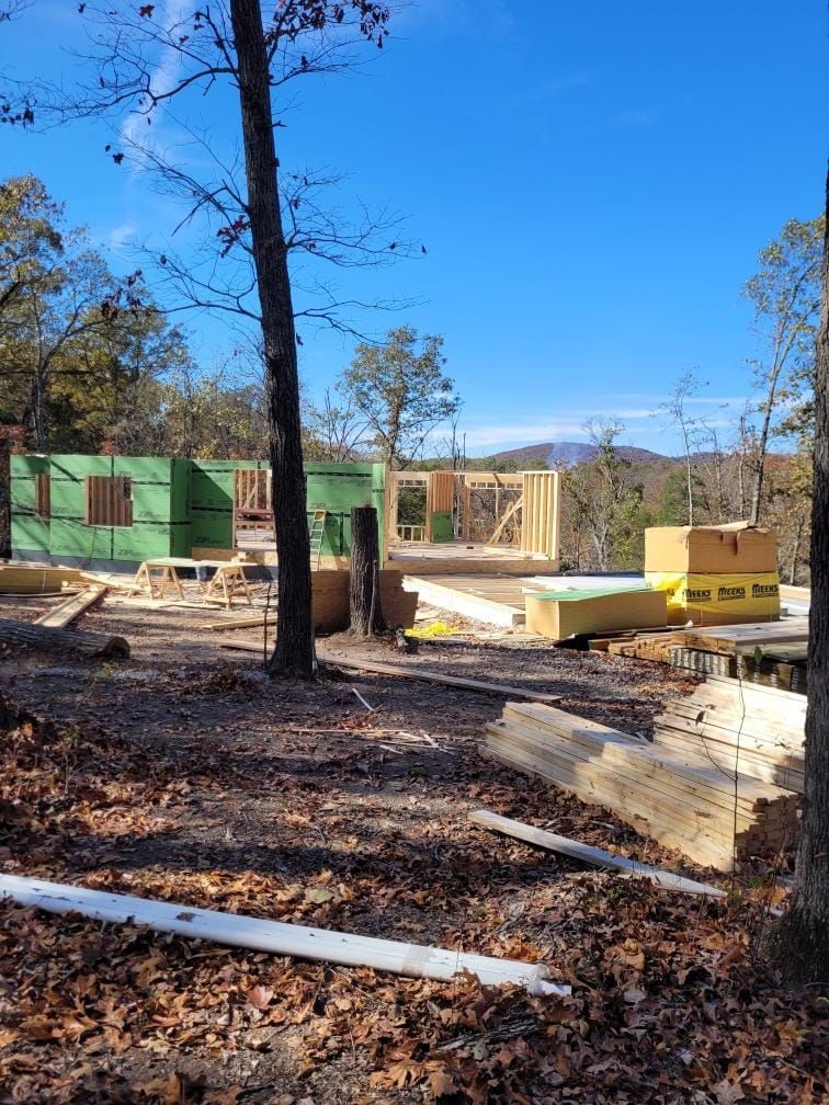 Construction of a wooden house frame on a wooded lot under a blue sky.