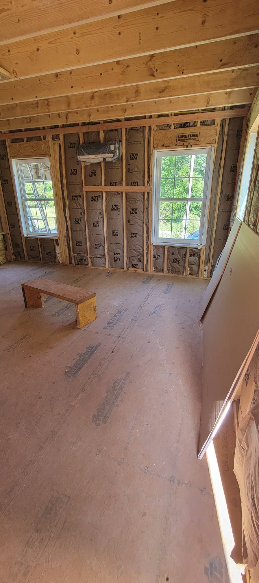 Interior of a room under construction, with exposed wooden beams, insulation, and windows. A wooden bench sits on the floor.