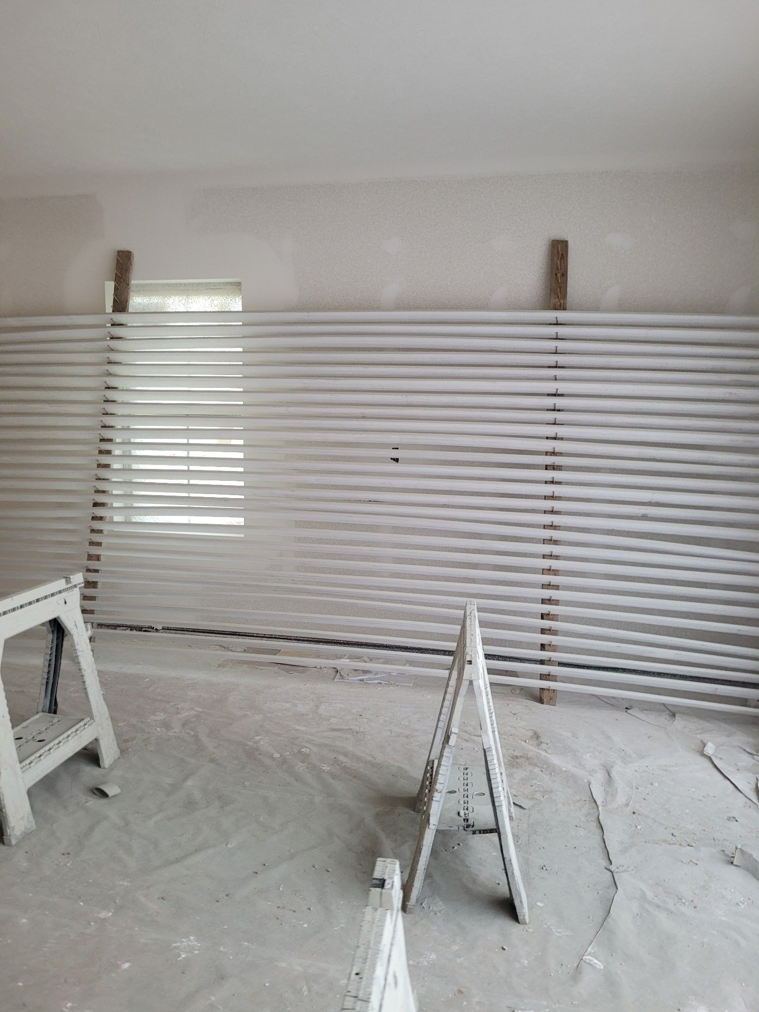 Interior room with a wall of white slats, partially covering a window. Two sawhorses are in the foreground.