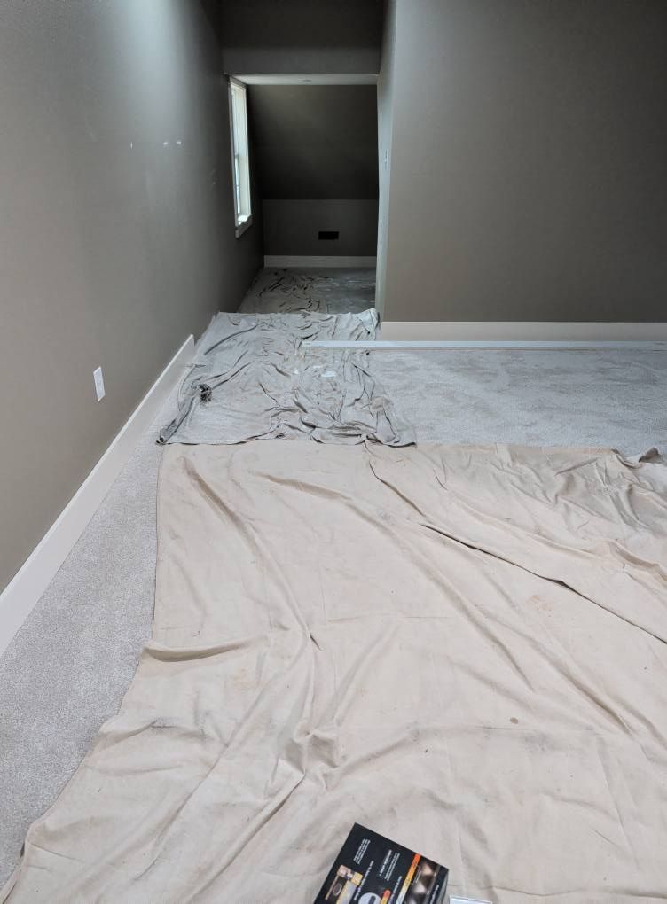 Room with gray walls, white trim, carpet, and a hallway. Paint cloth covers the floor.