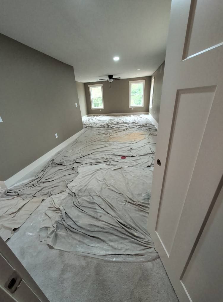 A room being renovated. Tan carpet covered in protective cloth. Gray walls and white trim. Two windows at far end.