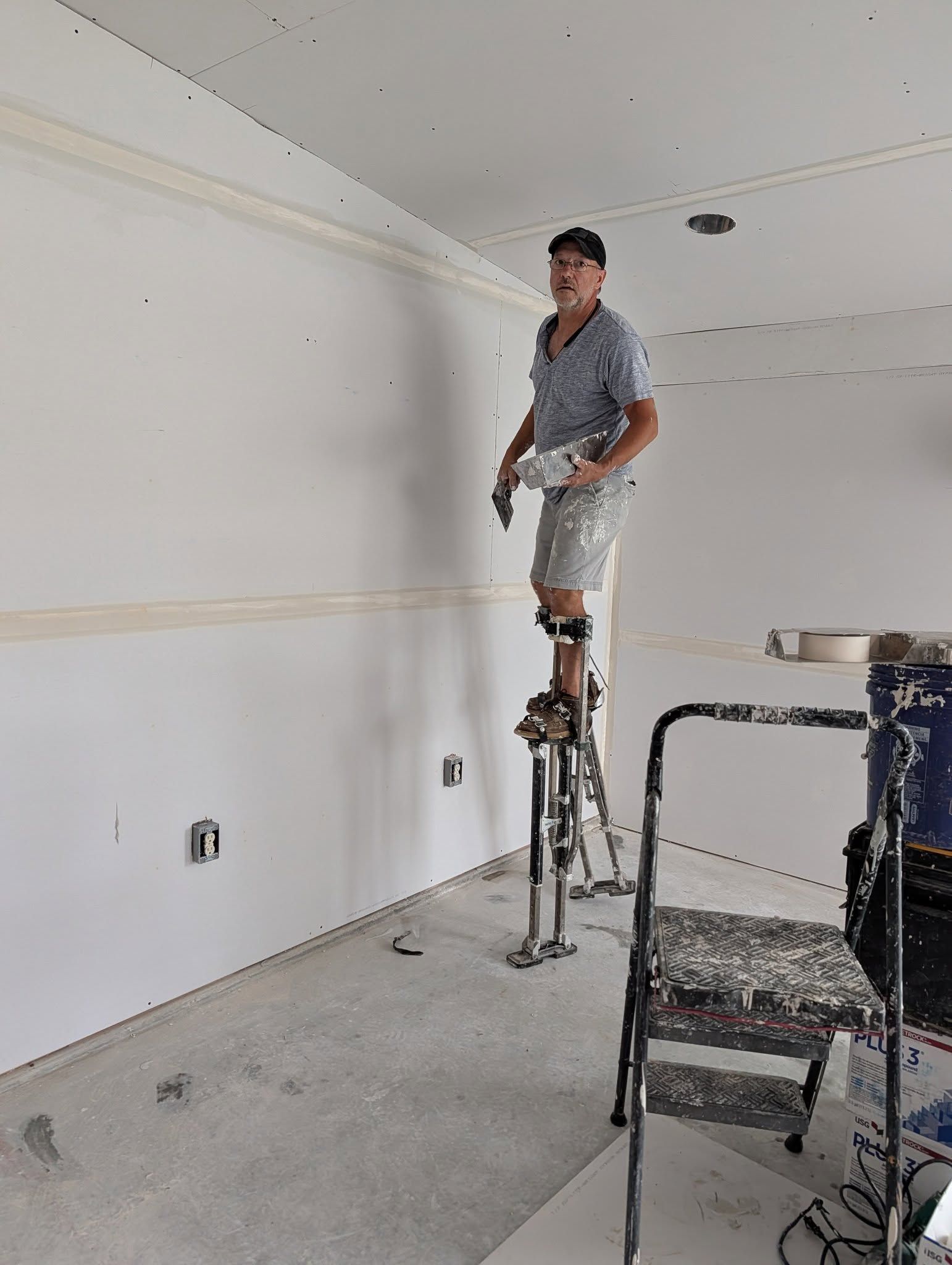 Person on stilts working on a wall, holding tools, in a room under construction.