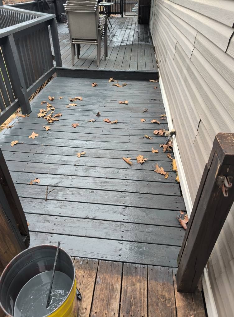 Outdoor deck painted gray with a bucket of paint, leaves scattered on the surface, and a building on the right.