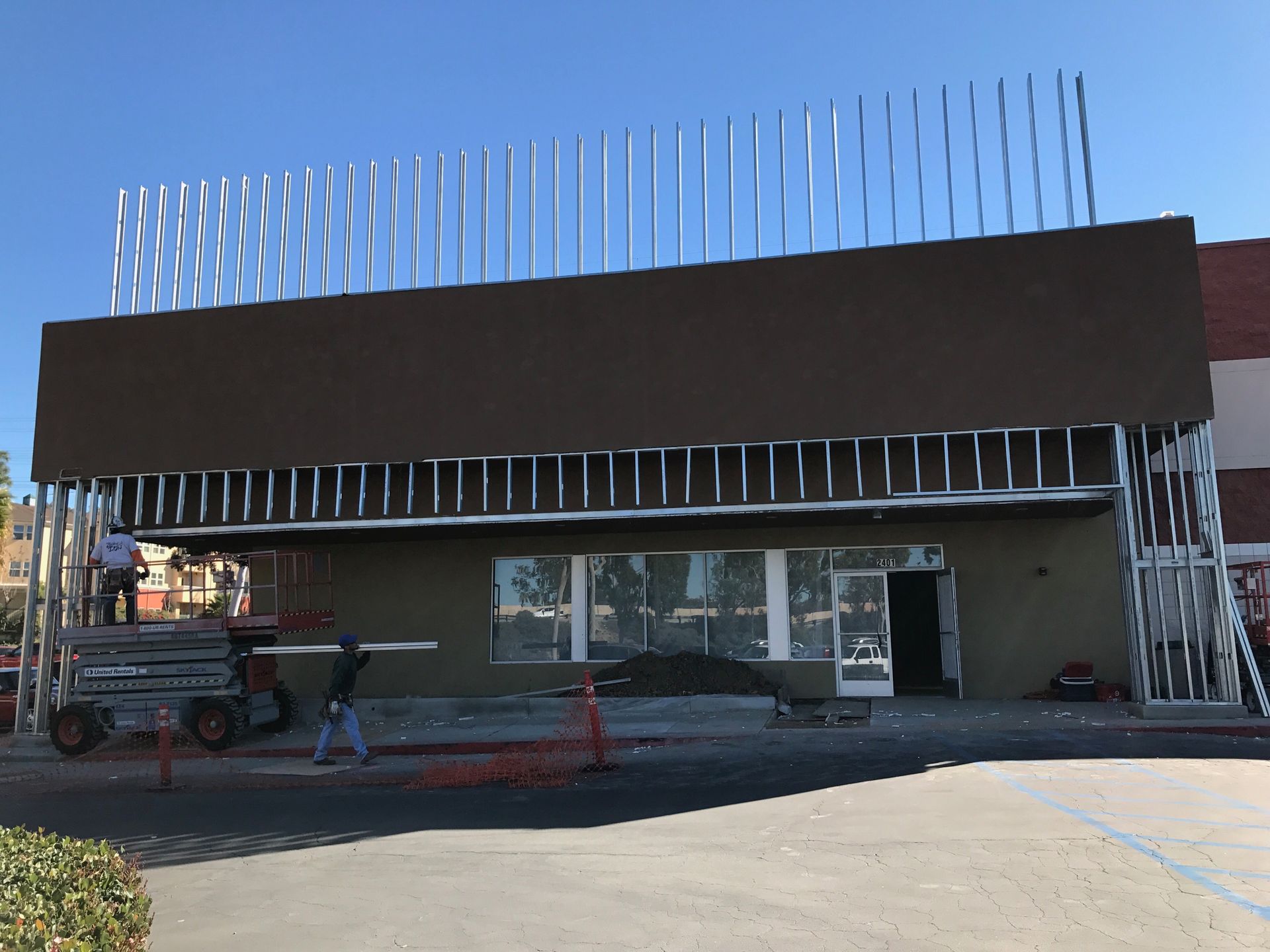 Building under construction, brown facade, silver metal framework, worker on lift, blue sky.