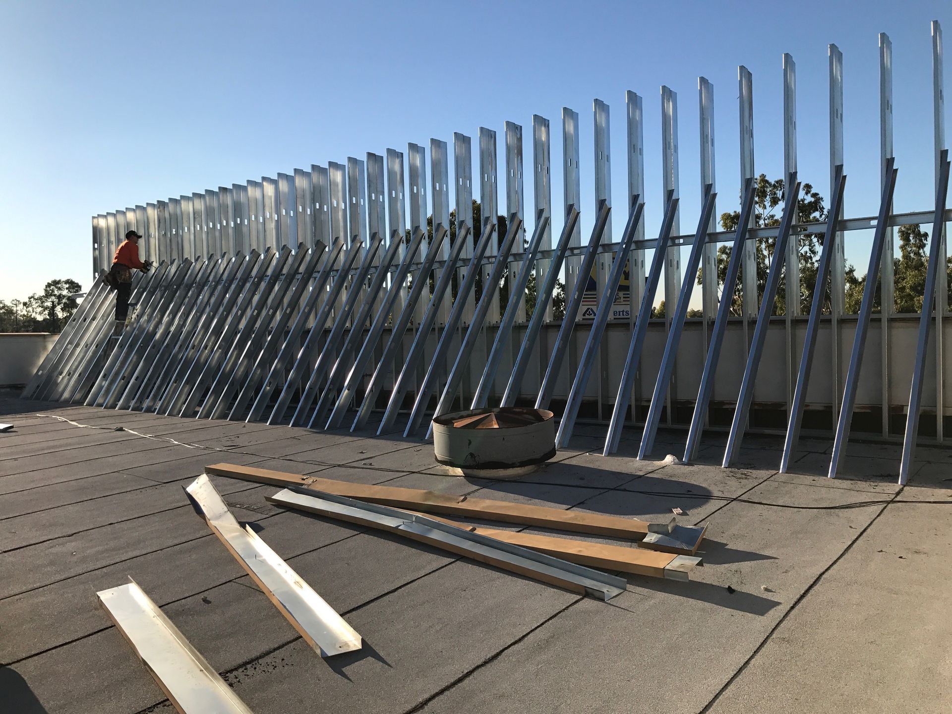 Construction of metal beams on a flat rooftop. A worker stands near the partially built structure on a sunny day.