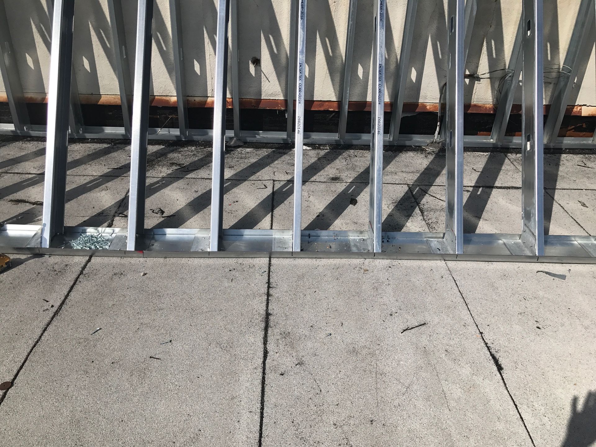 Metal framing on a concrete surface, with shadows cast across the ground, and a brown wall in the background.
