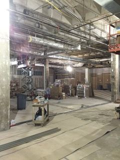 Interior of a construction site. Exposed pipes and wiring run along the ceiling. Drywall, tools, and materials are scattered.