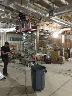 Construction site: worker on lift near ceiling, another on floor. Gray dumpster, exposed wires and ducts.