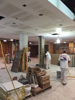 Construction site: Workers in white coveralls, tools, materials, unfinished ceiling, concrete floor.