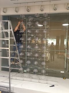 Man on ladder installing a transparent display with many dome-shaped objects. Interior setting, fluorescent lights.