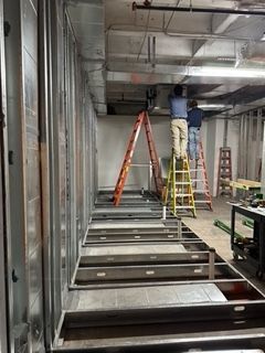 Construction workers on ladders installing ceiling fixtures, interior framing visible.