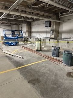 Construction site with a lift, caution tape, and copper pipes on the floor. Concrete walls and ceiling.