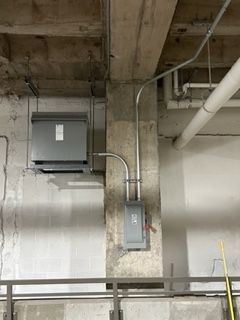 Electrical equipment mounted on a concrete wall. Includes a grey transformer and a panel box with conduit.
