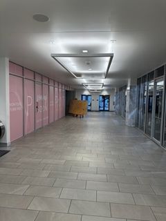 Hallway with rectangular ceiling lights, pink wall with lettering, gray tiled floor, and glass door at the end.