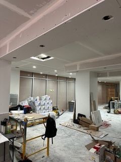 Interior construction site. Drywall and materials on floor, unfinished ceiling with recessed lights, windows.