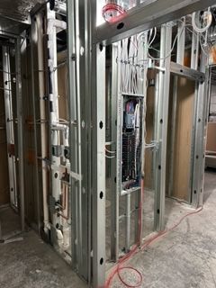 Metal framing of a wall with electrical panel and plumbing visible during construction. Concrete floor.