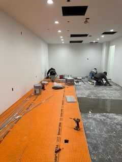 Workers installing tile flooring in a room. Orange underlayment, tools, and materials visible.