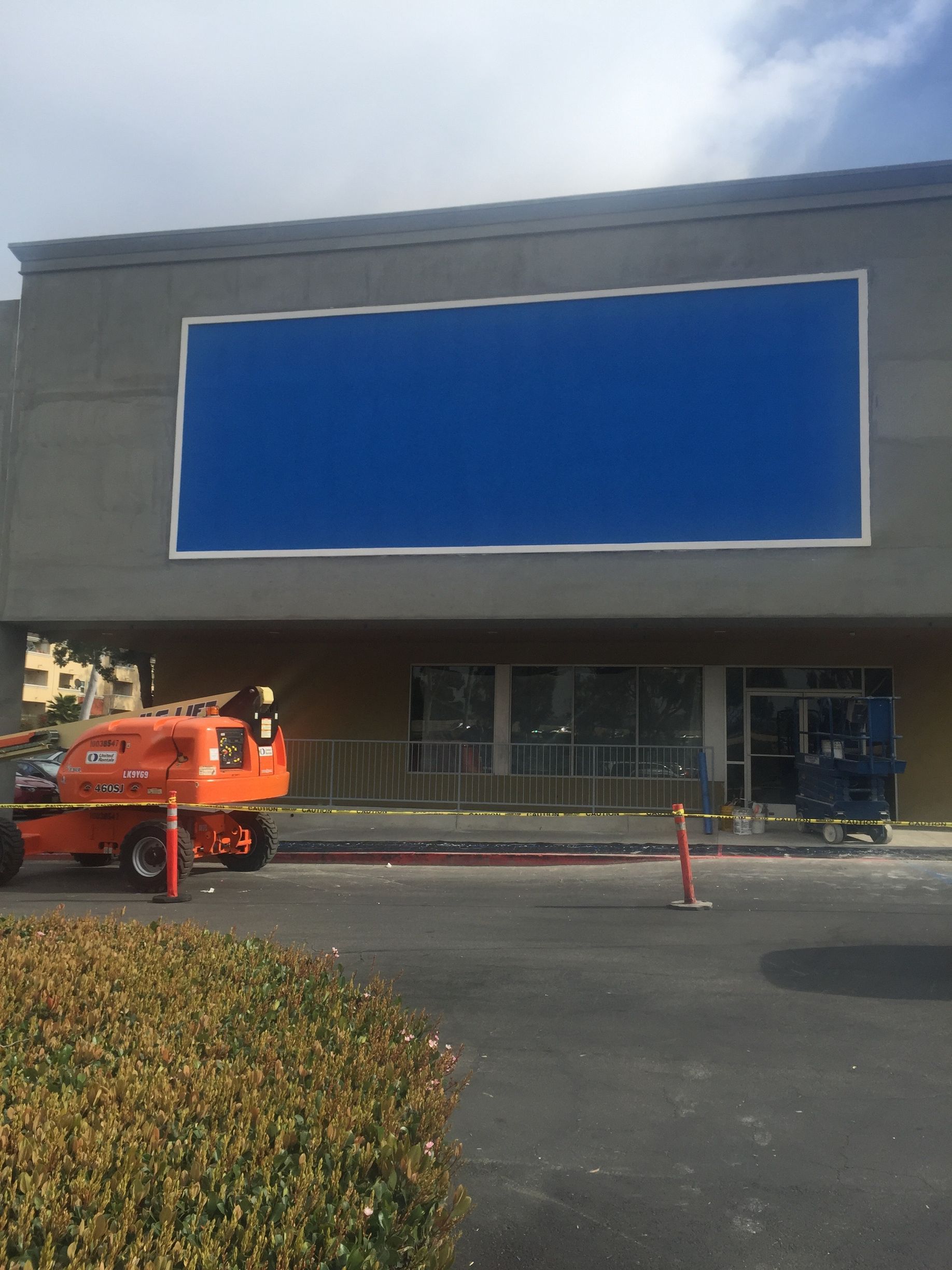 Exterior of a building under construction, with a large blue sign, scaffolding, and an orange lift.