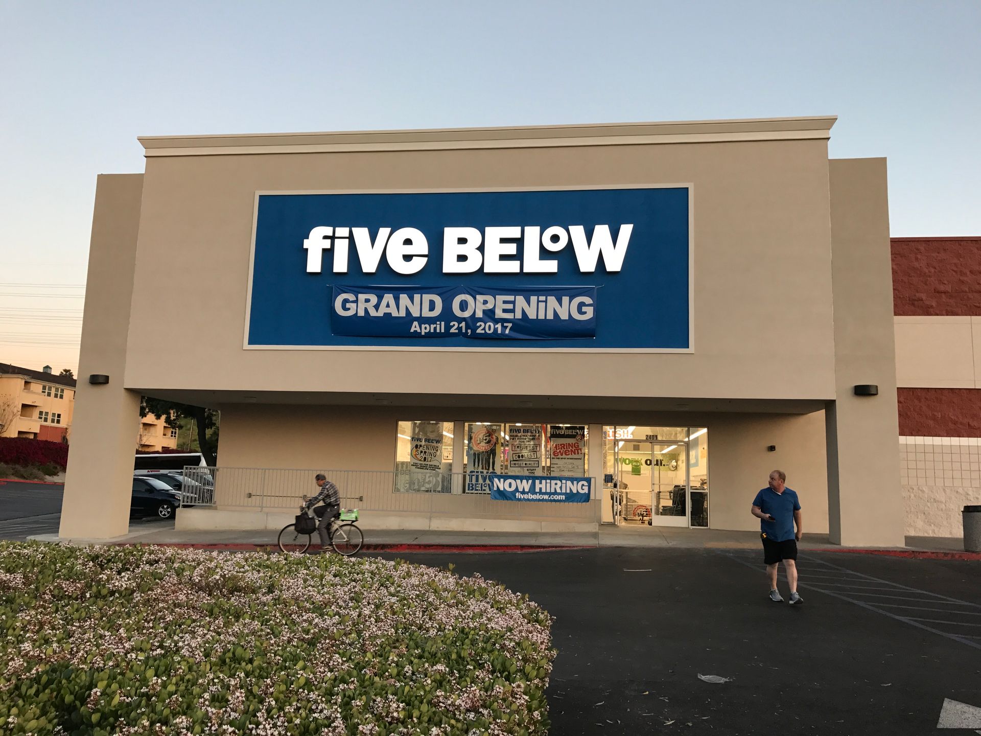 Five Below store with Grand Opening sign. Person on bike, another standing. Beige building, blue sign.
