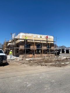 Building construction site; workers on scaffolding, blue sky. Brick and wooden framing visible.