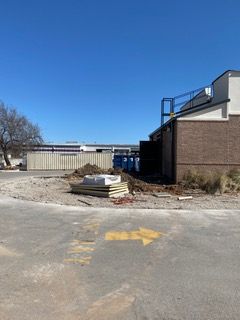 Construction site with building; dirt, materials, and a yellow arrow on asphalt. Blue sky.