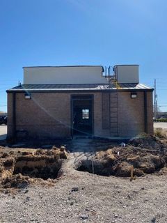 A brick building with an open doorway under construction, surrounded by dirt and gravel; a ladder runs up the side.