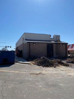 Brick building under construction on a sunny day. Dirt pile, blue dumpster, and road visible.