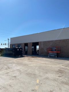 Exterior of a brick building with open garage doors; a dumpster and lift are visible on a concrete surface under a blue sky.