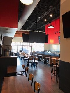 Interior view of a restaurant. Tables and chairs on light wood floors, red and black accents, TVs mounted on the ceiling.