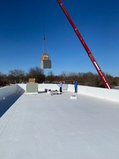 Crane lifting HVAC unit onto a white flat roof with two workers present on a sunny day.
