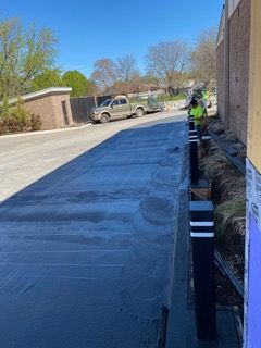 Construction site with wet concrete, black bollards, and a worker in a neon vest; truck in the background.