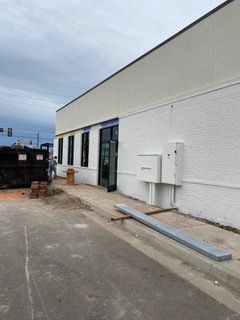 Exterior of a commercial building under construction; white walls, black windows, a dumpster, and a partially paved sidewalk.