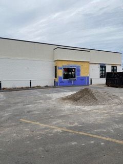 Exterior of a building under construction, drive-thru window framed with blue and yellow, gravel pile in foreground.