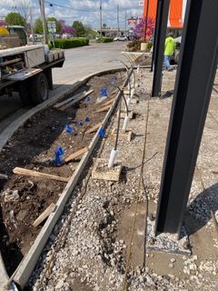 Construction site, curb and sidewalk. Blue stakes, wooden forms, dirt, gravel, and metal beams are visible.