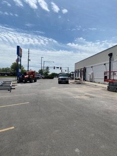 An empty parking lot with a white building on the right and a street with vehicles in the distance under a blue sky.