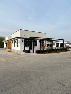 Modern building with black pergola and outdoor seating; white and tan exterior; on a paved lot.