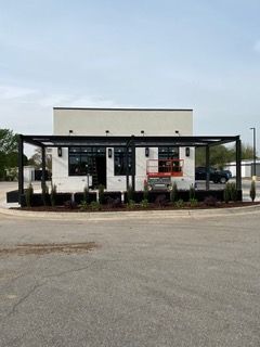 Building under construction, light brick exterior, black pergola, landscaping, parking lot.