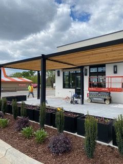 Exterior of a commercial building with a black pergola, planters, and a worker. Cloudy sky.
