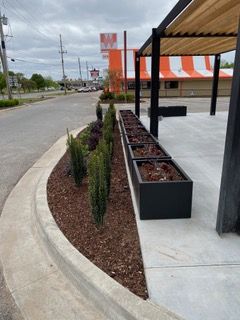 Planter boxes and shrubbery border a sidewalk near a building with an orange and white striped facade.