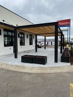 Outdoor dining area with a retractable pergola. Black planters and support beams, light-colored retractable cover, next to a building.
