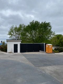 White building with a black door, black fence, and a portable toilet on a concrete lot, with trees in the background.