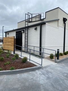 Building with a wheelchair ramp, ladder to the roof, and wooden fence. The building is white and black with a gray ramp.