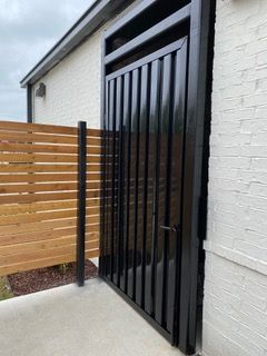 Black metal gate with vertical slats beside a wooden fence and white brick wall.