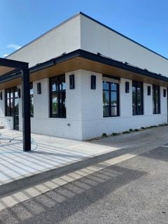 White brick building with black trim, windows, and pergola. Concrete sidewalk and driveway.