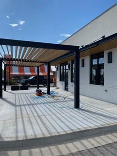 Pergola with slatted roof casting linear shadows on a concrete patio next to a white building.