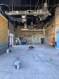 Interior of a building under renovation, showing exposed ceiling fixtures, concrete floors, and workers.