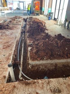 Construction site: trench with PVC pipes, dirt piles, and workers in the background.