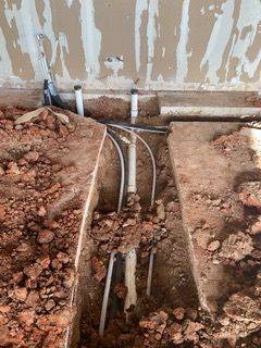 Open trench in red soil revealing plumbing pipes; unfinished wall above.