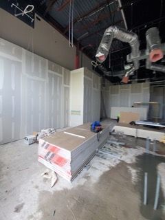 Construction site interior with drywall stacked on floor; exposed ceiling ductwork. Concrete floor, unfinished walls.