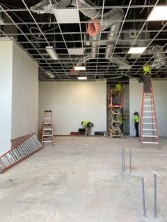 Construction site: Workers installing ceiling grid, electrical, and drywall. Ladders, lift, and concrete floor visible.